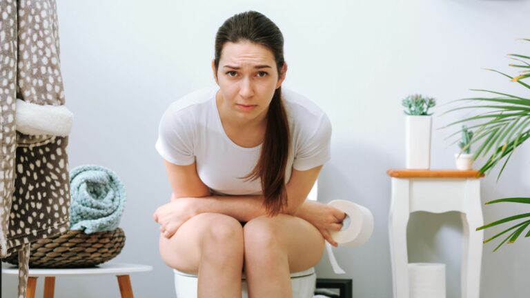 Femme assise sur les toilettes souffrant de constipation avec des douleurs au ventre et un rouleau de papier toilette à la main